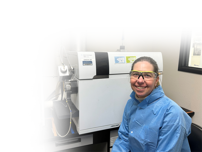 Smiling Nitto Avecia scientist in a blue lab gown and safety glasses standing beside an Agilent Technologies 7800 ICP-MS instrument used for elemental impurity analysis.